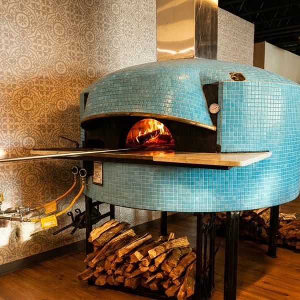 A blue tiled pizza oven with a roaring fire inside, surrounded by stacked firewood.