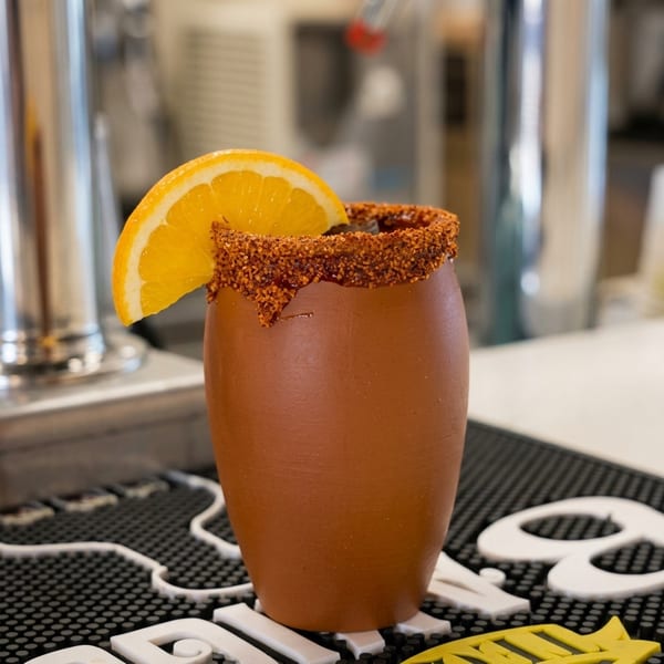 Orange cocktail in a clay-style glass with a chili-salt rim and orange wedge garnish on a bar counter.