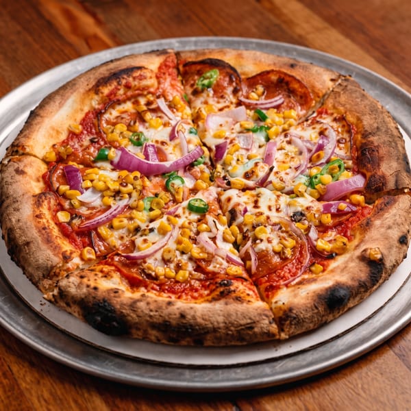 Wood-fired pizza topped with melted cheese, corn, red onion, green peppers, mushrooms, and tomato sauce on a metal plate.
