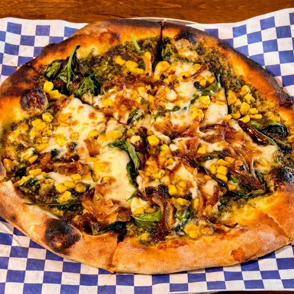 A sliced pizza with corn, spinach, caramelized onions, and cheese on blue and white checkered paper.