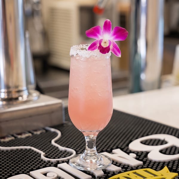 Pink cocktail in a stemmed glass, rimmed with sugar and garnished with a vivid purple orchid at a bar.